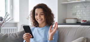 Woman with curly, dark hair in a light blue sweater is sitting in a beige sofa holding a black mobilephone. She ias waiving to the phone.
