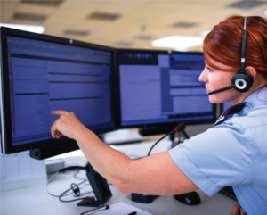 A red haired woman is wearing headphones and a blue uniform. She is inftont of a computer, pointing at the screen. She is working in a call center.